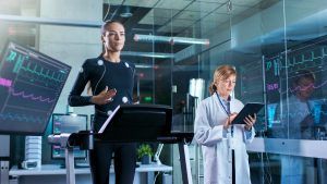 Woman Athlete Walks on a Treadmill with Electrodes Attached to Her Body while Scientist Holding Tablet Computer Supervises whole Process In the Background Laboratory with Monitors Showing EKG Readings